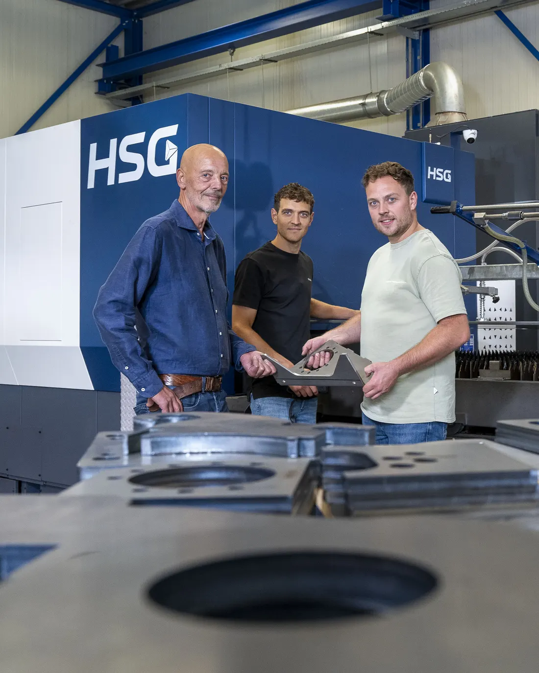 Hans, Koen en Tom Geurts houden samen een gesneden stalen onderdeel vast, voor de lasersnijmachine in de werkplaats.